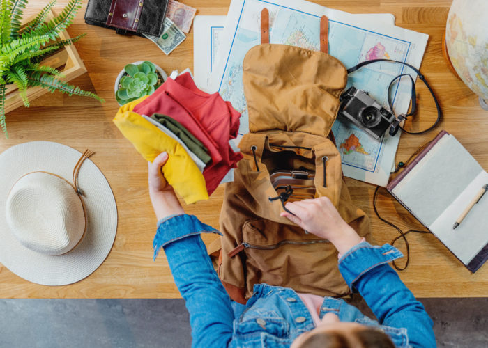 Aerial view of person packing backpack surrounded by maps, a camera, a globe, and a hat